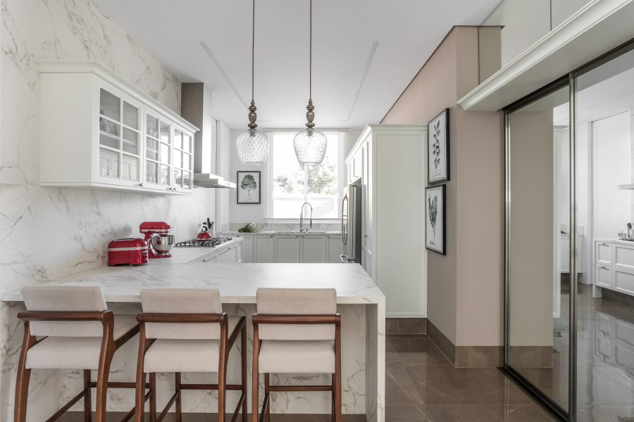 cozinha possui marcenaria em estilo clássico, com ampla iluminação natural e composição com texturas marmorizadas na bancada e em uma das paredes