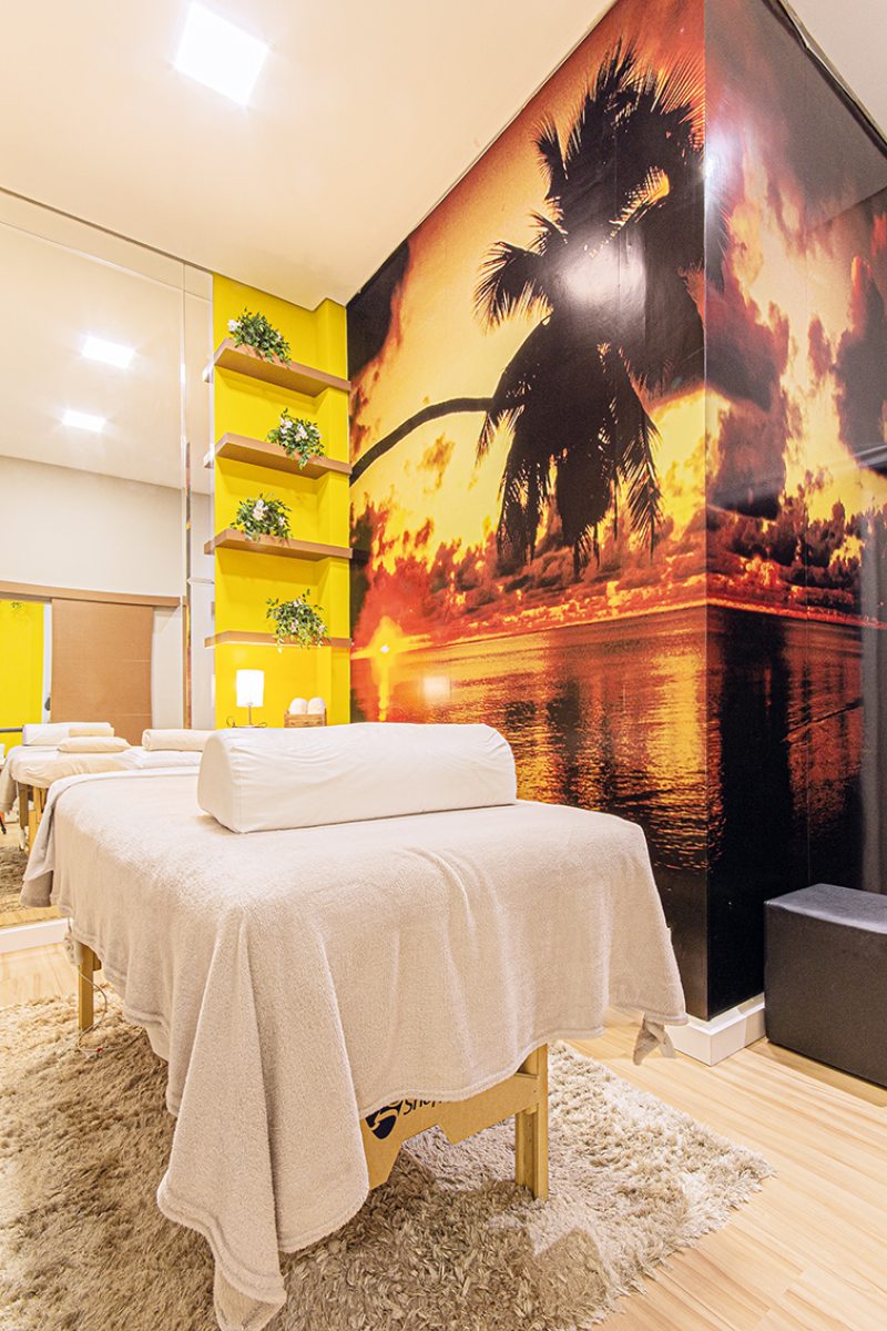Na sala de massagem, a profissional plotou uma paisagem paradisíaca em uma das paredes. Nichos em MDF amarelo com prateleiras amadeiradas completam o espaço