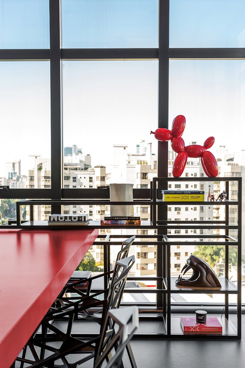 A mesa vermelha em frente ao grande pano de vidro é apoiada em uma estante de estrutura metálica repleta de livros de design e objetos assinados, como a escultura “Balloon Dog”, de Jeff Koons