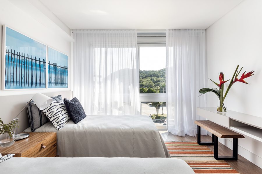 Em uma das suítes, tapete Kilim, cortinas leves que emolduram a vista para a natureza e marcenaria minimalista, com escrivaninha, banco e criado-mudo amadeirado