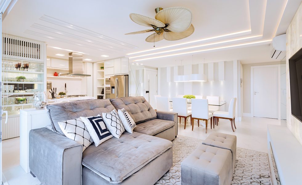 A porta principal do apartamento dá acesso ao espaço social integrado, composto por sala de TV, jantar e cozinha. O sofá em veludo cinza ganhou almofadas em tons off-white (Entre Sedas) que compõem com a base de cores neutras do projeto. Os recortes no forro de gesso foram iluminados com perfis lineares de LED (Grey House Iluminação), para um cenário acolhedor e moderno A porta principal do apartamento dá acesso ao espaço social integrado, composto por sala de TV, jantar e cozinha. O sofá em veludo cinza ganhou almofadas em tons off-white (Entre Sedas) que compõem com a base de cores neutras do projeto. Os recortes no forro de gesso foram iluminados com perfis lineares de LED (Grey House Iluminação), para um cenário acolhedor e moderno