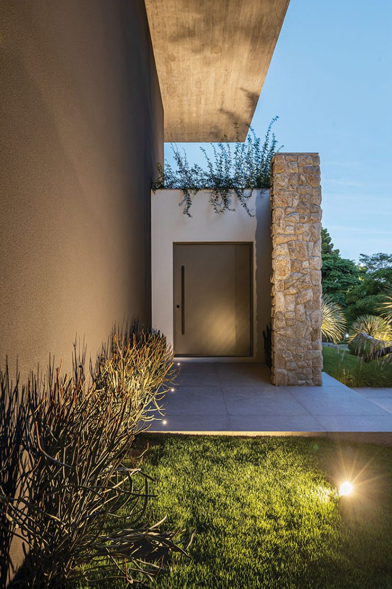 O volume de pedra natural camufla a grande porta de entrada principal executada em ACM na cor cinza. Da iluminação à paleta de cores, todos os  elementos decorativos foram escolhidos tendo os materiais construtivos elementares  como ponto de partida