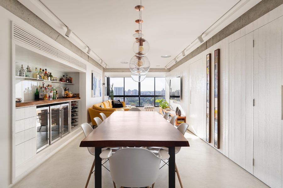 A sala de jantar integra a sacada gourmet e o estar íntimo, com a mesa em madeira natural de bordas irregulares. O destaque do ambiente fica  por conta do bar embutido, com atendimento de refrigeração para os diversos tipos de bebidas, espaço para cristaleira e preparo de drinks