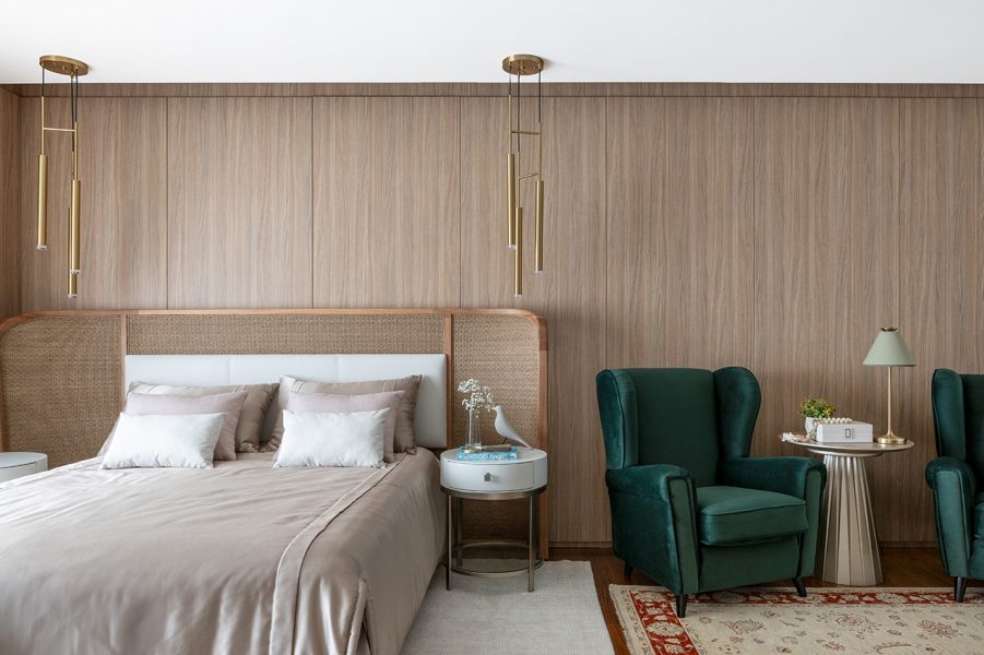 Na suíte principal, a cabeceira estofada com detalhe em palhinha compõe com os tons de bege e marrom claro das roupas de cama. Poltronas verdes e mesa lateral da Sierra formam o canto de leitura, enquanto painéis amadeirados revestem a parede principal. Tapete com estampa  discreta e luminárias pendentes douradas completam o ambiente.