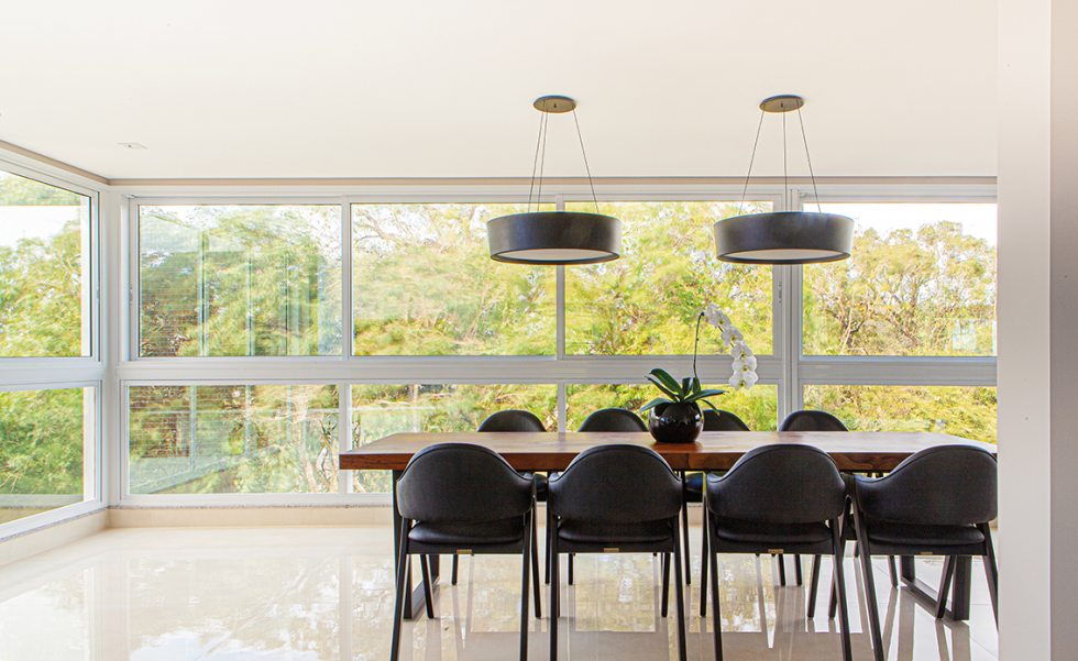 Integrada ao espaço gourmet, a sala de jantar possui vista para a natureza exuberante do entorno. A mesa de madeira de demolição e o mobiliário de cores escuras contrasta com a ampla luminosidade natural