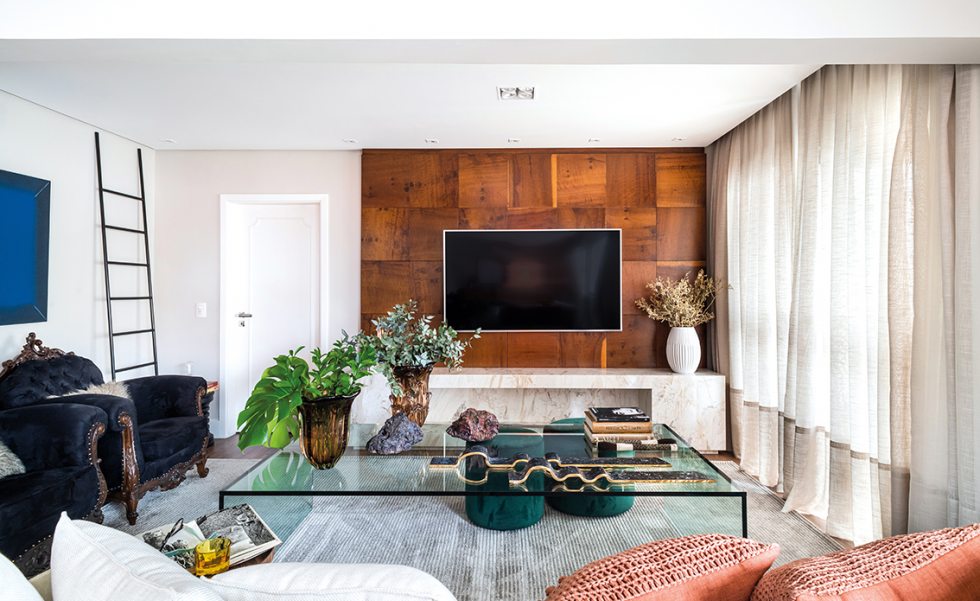 A sala de TV recebeu painel de madeira natural de imbuia, proveniente do retrofit dos antigos armários do apartamento. Os quadrados de 45 centímetros foram alinhados em sentido horizontal e vertical, sem que os veios ficassem necessariamente alinhados. Abaixo, encontra-se uma moldura linear em mármore Branco Paraná para uma lareira a álcool. O sofá foi escolhido a dedo na Momenttum