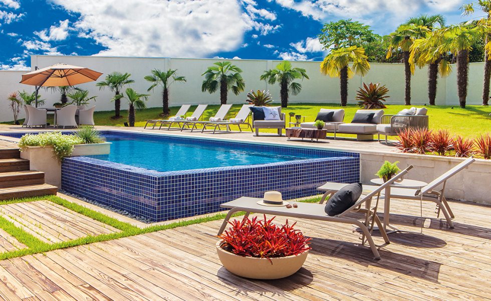 Nos fundos da casa, área externa com espreguiçadeiras, mesas, sofá, poltronas, mesa de centro e ombrelone todos da Green House, em tecidos e acabamentos resistentes a intempéries Nos fundos da casa, área externa com espreguiçadeiras, mesas, sofá, poltronas, mesa de centro e ombrelone todos da Green House, em tecidos e acabamentos resistentes a intempéries