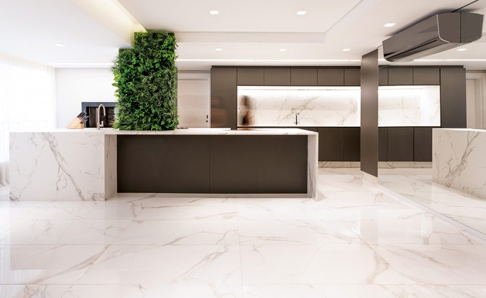 Após a remoção da parede que dividia a sala da cozinha, o pilar permanente foi incorporado à extensa ilha de quatro metros em Dekton Aura, com efeito marmorizado que combina com o porcelanato do piso. As três faces do pilar foram revestidas com espelhos, para mimetizá-lo, e a face voltada para o living recebeu vegetação natural
