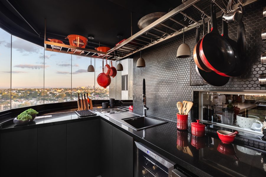 A cozinha se destaca como um contraponto ao ambiente predominantemente branco do apartamento, criando um efeito de contraste marcante. O uso de tons escuros e acabamentos em preto nos armários e bancadas confere um visual sofisticado exaltado pelo revestimento hexagonal das paredes que adiciona textura e interesse visual, quebrando a monotonia que poderia surgir em um espaço totalmente neutro