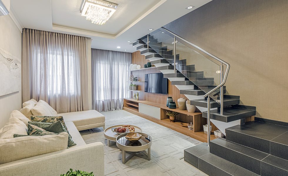 A sala de estar ganhou móvel de tV projetado para acompanhar o desenho da escada, com guarda-corpo de vidro. O lustre decorativo com cristais, no centro, proporciona um cenário acolhedor, em conjunto com o tapete de textura suave e o sofá em tom off-white