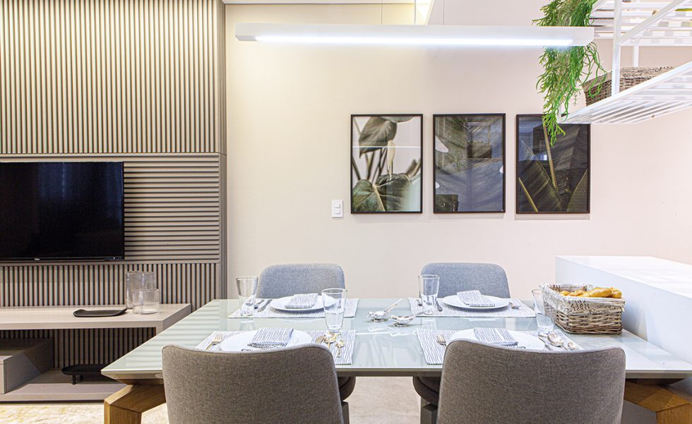 A mesa da sala de jantar é ladeada pelo balcão da cozinha, para maior otimização de espaço. A TV foi posicionada sobre o painel ripado da Todeschini Batel na cor Nuage, permitindo sua visualização tanto da sala quanto da cozinha e jantar