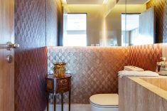 Neutro, o lavabo tem piso e bancada de mármore travertino navona e recebe um papel de parede de palha bronze com desenhos feitos a laser