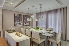 A sala de jantar traz cortinas com detalhes no barrado em tela e mesa de jantar com oito lugares, adquiridos na M. Decor. Sobre a mesa, seis pendentes espelhados posicionados em tamanhos e comprimentos diferentes para criar movimento. A parede dos fundos recebeu papel de parede em tonalidade off-white (M. Decor)