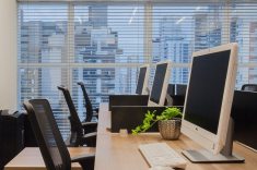 A mesa de trabalho possui pés em aço preto e tampo em madeira. As divisórias contam com persianas que podem ser utilizadas para isolar ou integrar espaços, conforme a necessidade.