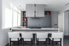 A cozinha é planejada pela Florense traz tampo em quartzstone Royale Blanc. Nos armários e gavetas, laminado Rovere Argento, espelhos fumê Acidato e puxadores Tech (Florense). A coifa dupla cria uma volumetria de impacto visual. O piso em quartzstone cinza foi escolhido pela resistência e efeito visual. Nas janelas, persiana Rolô Panamá (Luxaflex)